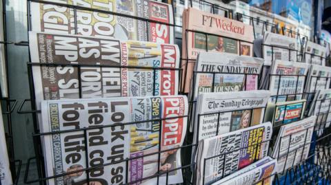 A rack of national newspapers seen from a side angle, including the Mirror, the Sunday People and the Sun on Sunday at the front left of the picture.