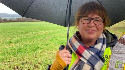 Loraine Hopkinson, who has brown hair and eyes, and wearing yellow coat with checked scarf, is standing under an umbrella in a field with hills in the background. She is smiling.