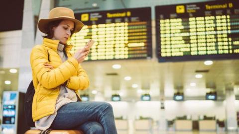 A lady in a hat and yellow jacket sat in front of airport departure boards, sat on her suitcase and looking in to her mobile phone.