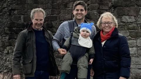 A family group standing outdoors in front of the remains of an old stone wall, part of an historic ruin or castle. The people are dressed in outdoor clothing, including jackets and boots, and the man in the centre is carrying a small child. Behind the group there is a scenic view of green fields stretching into the distance under a partly cloudy sky.