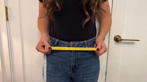 A woman in jeans stands with a yellow tape measure across her waist.