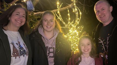 A family of four, mum, dad and two daughters, wearing Christmas jumpers, smiling at the camera, standing in front of a large illuminated reindeer decoration.