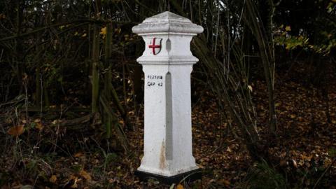 A white post with a pointed top with an England flag on the front surrounded by trees