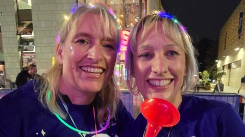 Andrea Corkhill and her sister Lindsey Moore smiling for the camera. Both women have straight blonde shoulder-length hair and are wearing fairy lights on their heads and glowstick necklaces.