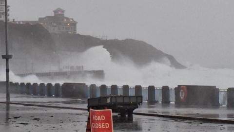 Waves crash onto the shore at Onchan Head, there is a road closed sign in the foreground and debris across the road.