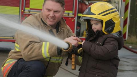 Benny, four years old, blasts a fire hose with the help of a real fireman beside him. A fire engine is visible behind them.