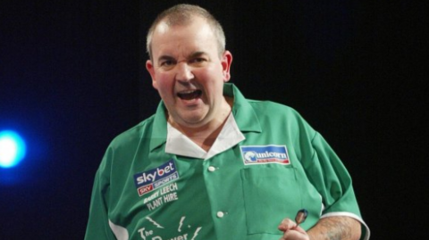 A man with short cropped hair is gripping a dart. He is wearing a green shirt with sponsor names on it