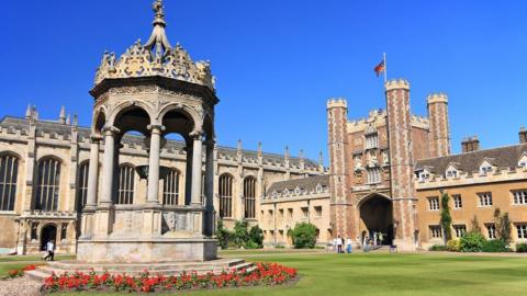 A court at Trinity College Cambridge has a manicured lawn. There are ornate buildings around the edge and people are walking around. In the foreground is a decorative stone fountain with red flowers planted around it