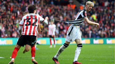 An action shot during Sunderland v Newcastle United in the 2024 FA Cup third round. Sunderland full back Trai Hume, wearing a red and white striped shirt with the number 32, has his back to the camera. In his right had he holds little more than a thread of winger Anthony Gordon's black and white striped shirt. That thread is tearing from the bottom of Gordon's shirt as he moves away from Hume. Other players and a section of crowd at the Stadium of Light are blurred in the background.