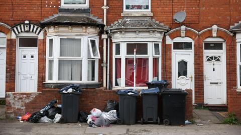 Several black bins, some overflowing, with piles of rubbish next to them, in front of a small wall. The wall is in front of a row of terraced houses.