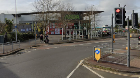 An Asda store is at a traffic lights junction in Battle Road, St Leonards. There are two bus shelters on the pavement near a bus layby. The Google image shows the road in daytime and there are people walking around.