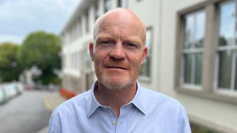 A man looking directly at the camera wearing a blue shirt. Background is blurred but a building on the right can be seen as can a road to its left. Trees and cloudy skies behind.
