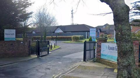 Entrance to Rowan Garth Care Home in Anfield, Liverpool. It shows black open gates into the car park for the building, with a number of welcome signs reading Rowan Garth Care Home. It is a dry day.