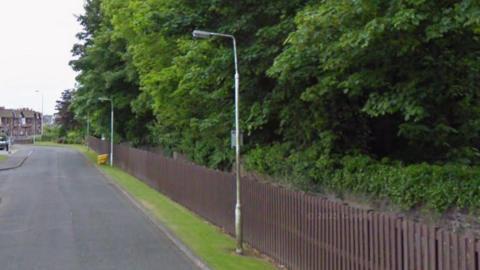 A road and green leafy wood are separated by a long brown wooden fence. There are lampposts and houses in the distance.