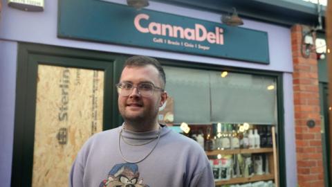 Tom Richards, owner of Canna Deli in Pontcanna, stands in front of his store. A boarded up door window can be seen in the background to the left. To his right, shelves of coffee and bottles can be seen in the window. The canna deli store sign is above his head in the background, in pink font with a navy background.