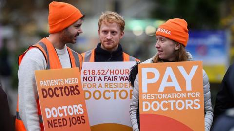Three resident doctors stand on a picket outside St Thomas' hospital in London at the start of a 5-day strike on 14 November 2025. They wear orange hats and hi-viz jackets and hold plackards reading "Patients need doctors, doctors need jobs" and "Pay restoration for doctors".