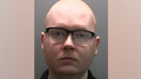 Mugshot of Daniel Carter who is bald and has a large forehead. He is wearing black-rimmed glasses and is looking at the camera impassively.