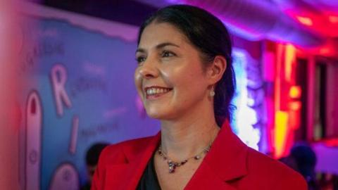 Llun yn dangos menyw efo gwallt yn tywyll a siaced coch yn edrych i ffwrdd o'r camera.