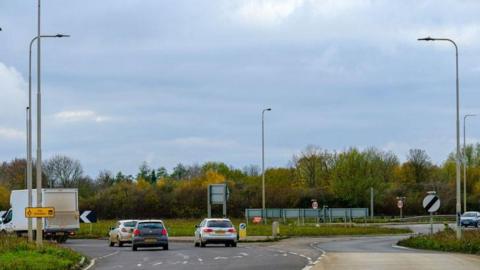 Image of the A16 round about with cars driving around it and street lamps can be seen on either side of the road.