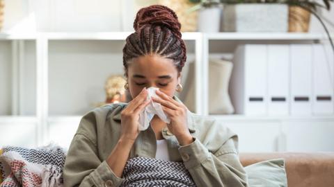 A woman inside in a green top blows her nose on a tissue while sitting on the sofa