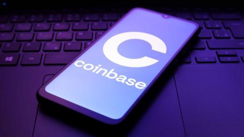 Coinbase's logo displayed on a smartphone screen. The handset is lying on top of an illuminated laptop keyboard.
