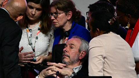 A bearded man, COP president Andre Correa do Lago lies back as several others surround him, looking worried, during a critical moment in the COP30 talks