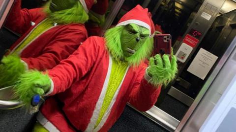 Jeff dressed as the Grinch with a green mask and fur, and a red and white Santa suit poses for a selfie