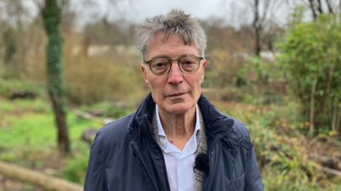 Dirk Lips has a blue jacket and an open necked white shirt. He is wearing glasses and is looking directly at the camera.