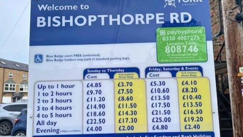 A blue and white parking board showing prices at Bishopthorpe Road car park. Up to one hour costs £4.85 and all-day parking costs £22.50 from Sunday to Thursday.