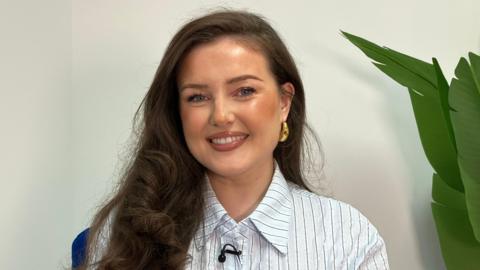 Mae Lauren Bell yn edrych yn syth ymlaen, mae ganddi wallt hir tywyll ac mae'n gwisgo crys golau gyda streipiau. Mae planhigyn gwyrdd wrth ei hochr.