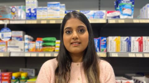 A woman with long brown hair wearing a pink knitted cardigan stands in front of boxes of medication.