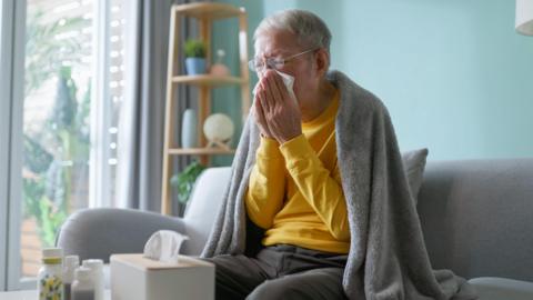 An older woman with grey hair and glasses blows her nose while sitting on a sofa with a grey blanket round her shoulders. There is a box of tissues on the table in front of her as well as various bottles that may be medicine.