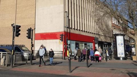 Roadside view of the TK maxx building -with a red entrance to a large cream building. People can bee seen walking in front of the building onto the road.
