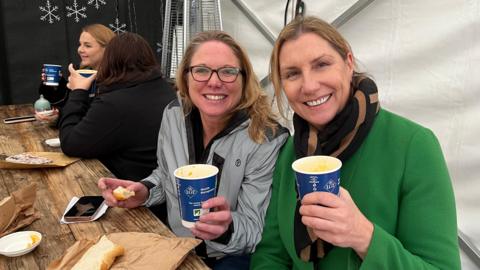 Liz Casely (Left) and Flo Highland (right) were among the happy customers at the annual event. Liz wears a reflective, silver waterproof jacket, while Flo is wearing a green coat and a black and beige scarf. Both have golden hair and are holding their paper cups of soup and smiling