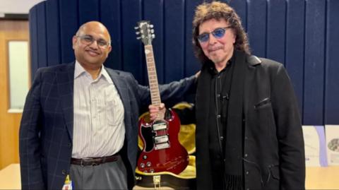 A bald headed man with glasses a white shirt and a blue jacked next to a man with black curly hair, blue-tinted sunglasses and a black coat with a dark brown guitar between them