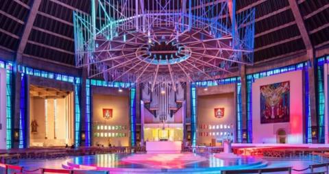 A large circular chandelier-style feature with a hole in the centre hangs from the ceiling, above a circular altar. Rows of wooden benches encircle the stage and the cathedral is lit in blue, pink, purple, pink and orange.