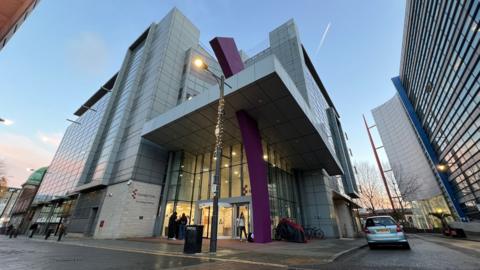 A large multi-storey modern building at the university's Southend campus. There are cars parked on the pavement outside too. It is evening time and it appears it has been raining.