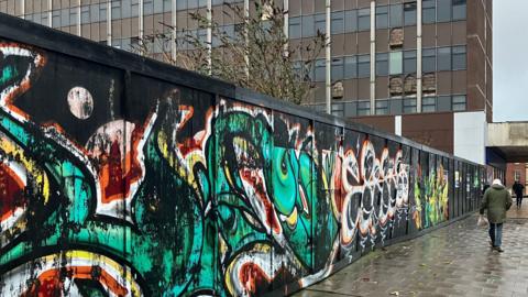 A hoarding surrounding a development site is covered in graffiti in Hanley in Stoke-on-Trent. A man holding a shopping bag walks past colourful green orange, white and yellow symbols on a black hoarding. High-rise buildings in a dilapidated state can be seen behind.