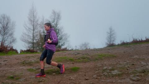 A woman with brown hair tied back is wearing a purple waterproof jacket, black three quarter length leggings and purple and pink trainers. She is running down a hill which is mostly brown earth with patches of green grass