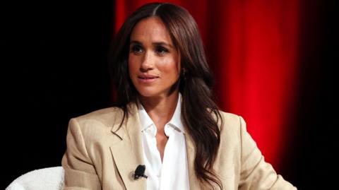 Meghan, the Duchess of Sussex sat down on a white chair wearing a beige suit and white shirt