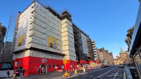 A white plastic sheet covering scaffolding around a hotel building Edinburgh. The sky above is blue. The lower half of the scaffold is covered by red, wooden panelling.