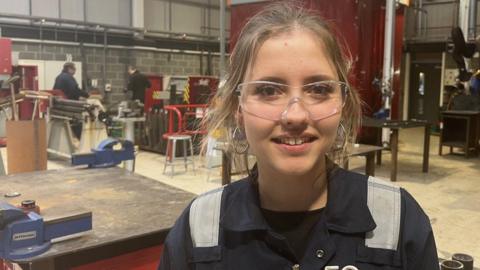 A young woman with long blond hair tied up smiles looking at the camera a she stands in a workshop. She is wearing blue coveralls and safety glasses. Two men are at a work table in the background behind her.