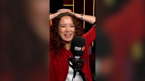 A woman, Laufey, with her hands on her head smiling in front of a mic. She wears a red cardigan