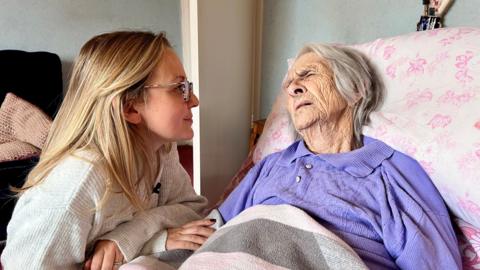 A 98-year-old woman in a purple top is lying in bed with a slightly pained expression on her face. Her granddaughter is kneeling next to the bed and holding her hand.