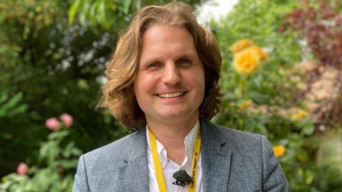 Steffan Aquarone is standing outside in front of green trees and bushes. He is looking directly at the camera and smiling. He is wearing a grey suit jacket and a white shirt.