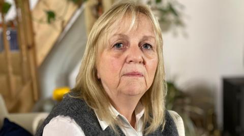 Nicky Jones is looking into the camera. She is wearing a white shirt and a knitted grey jumper. She has blonde hair. A TV, plant and staircase are blurred in the background.