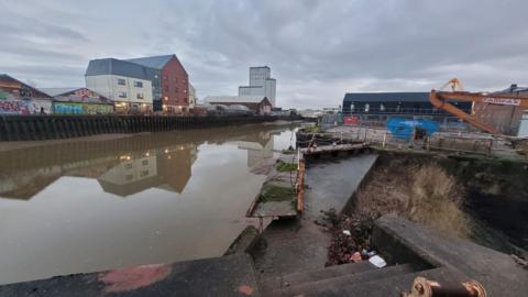 The photo show the rather industrial setting of the River Hull, with concrete steps and discarded rubbish. An old rusty and broken foot bridge is covered by moss. The water is dirty and brown.
