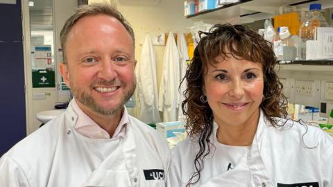 Two scientists, Professors Ed Wild and Sarah Tabrizi, are pictured from the shoulders up. They are both smiling and wearing white lab coats. Prof Wild on the left has a tightly cropped beard. Prof Tabrizi on the right has long wavy hair. They are clearly in a laboratory with scientific equipment on shelves in the background.
