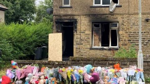 Tributes at the scene of the fire, including flowers next to the black burnt out windows and doors of the house
