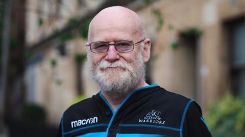 Person wearing a black Glasgow Warriors rugby shirt with blue stripes and Macron logo, standing outdoors in front of a stone building with greenery